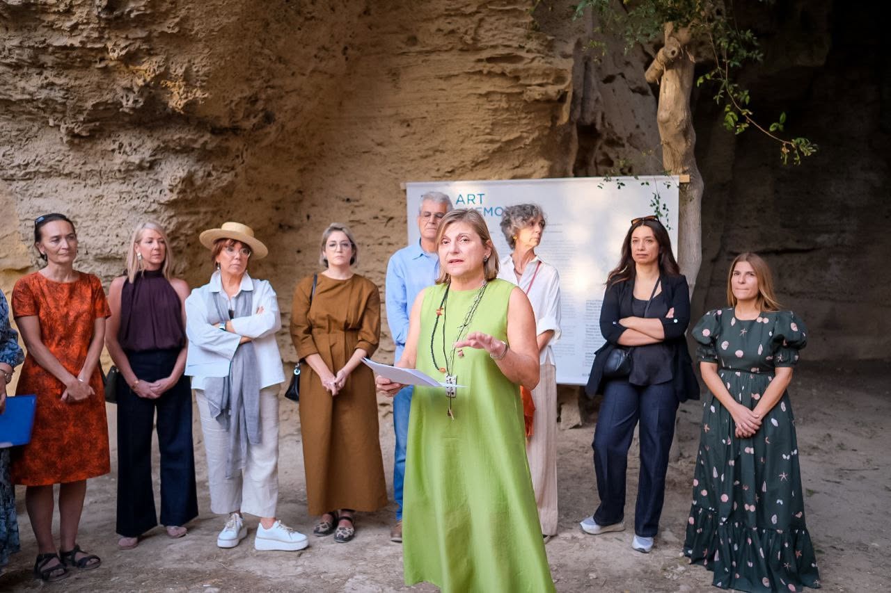 A speaker addresses a group of attendees gathered beneath a limestone rock face, with an information panel behind them