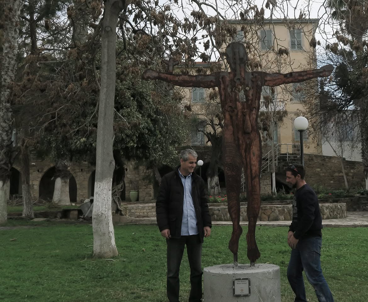 Two men stand beside a tall rust-coloured metal sculpture of a figure with outstretched arms in a garden