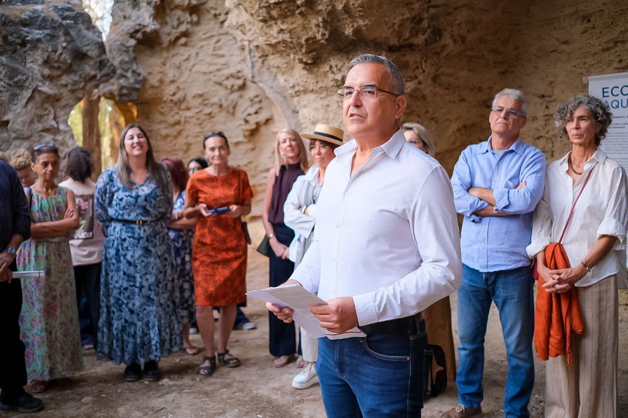 A man in a white shirt addresses attendees at an open-air gathering beside a rock wall
