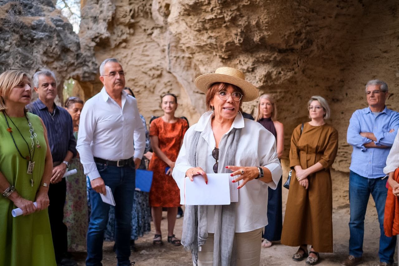 A woman in a straw hat speaks with notes in hand while attendees listen around her