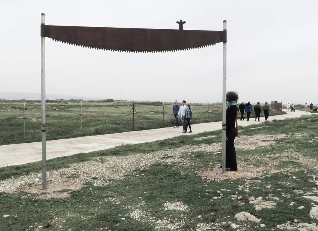 An outdoor installation of a large saw blade raised on two poles beside a coastal footpath